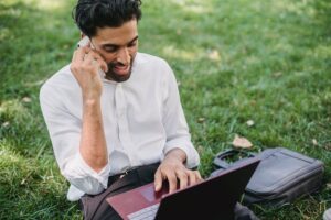 Man sitting on grass talking on the phone and using a laptop.