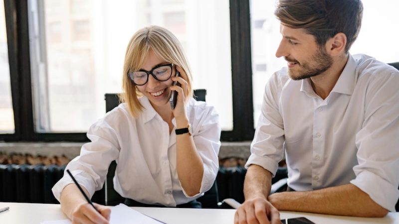 Two coworkers, one on the phone, in a sunny meeting room.