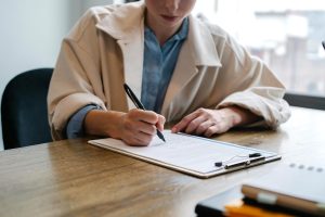 Person sitting at a table, next to a window, filling out an application on a clipboard.