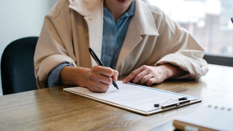 Person sitting at a table, next to a window, filling out an application on a clipboard.