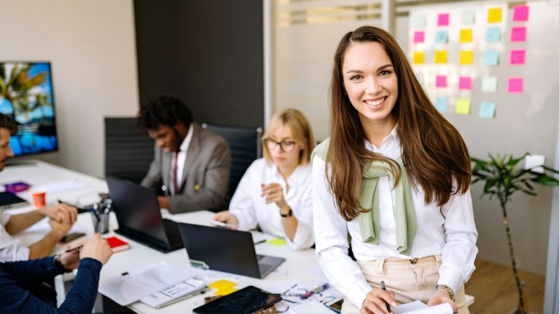 Office with people working on laptops, one woman smiling at the camera and holding a notepad.