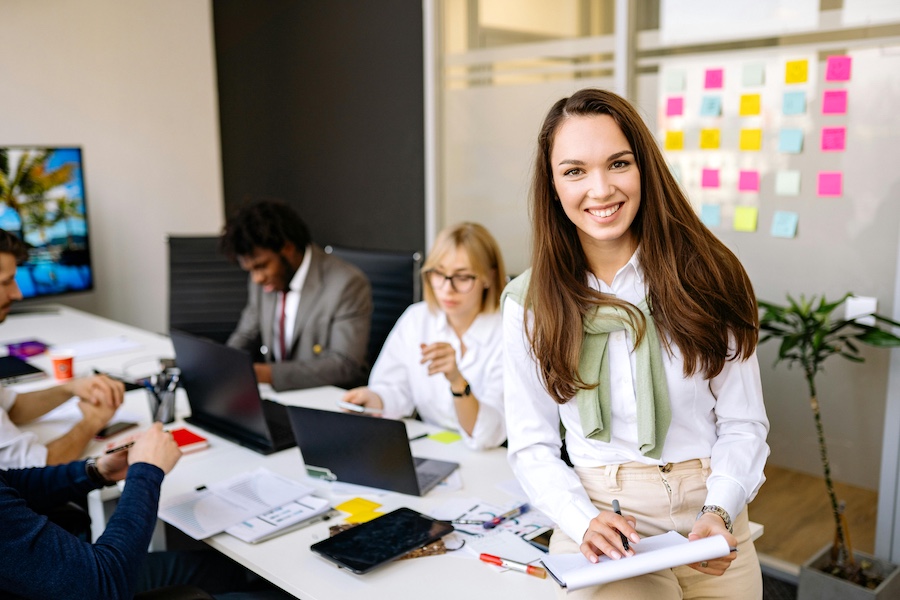 Office with people working on laptops, one woman smiling at the camera and holding a notepad.