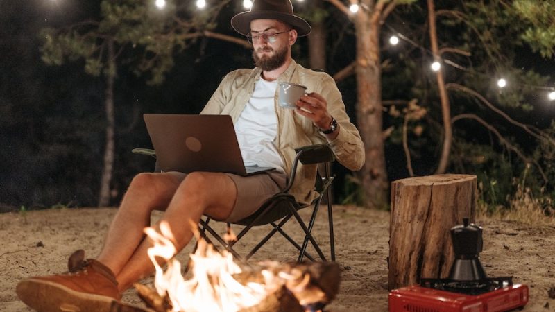 Man sitting in front of a campfire at a campsite with an open laptop.
