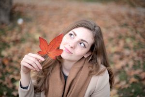 Woman outside in a coat looking up at a red leaf she is holding.