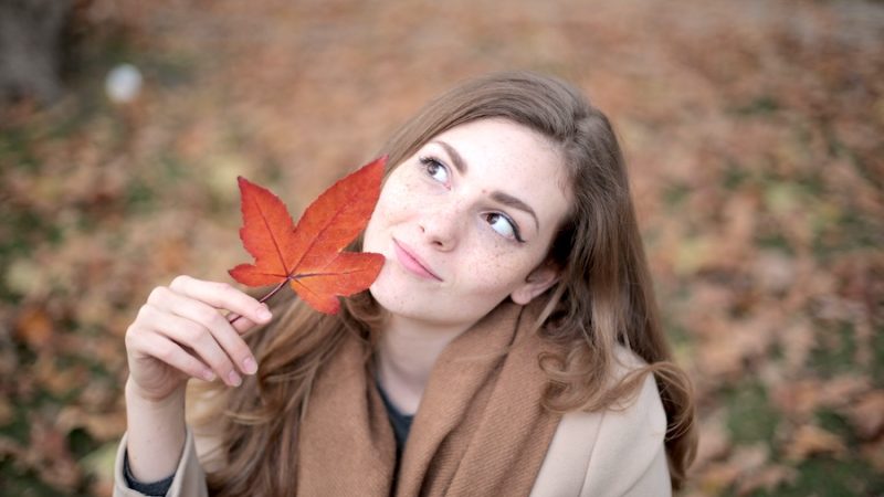 Woman outside in a coat looking up at a red leaf she is holding.