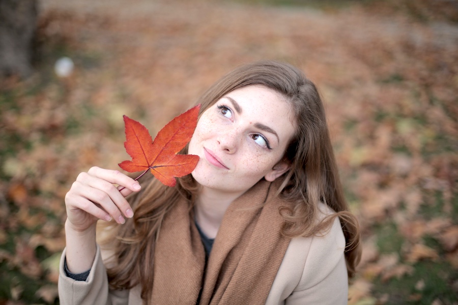Woman outside in a coat looking up at a red leaf she is holding.