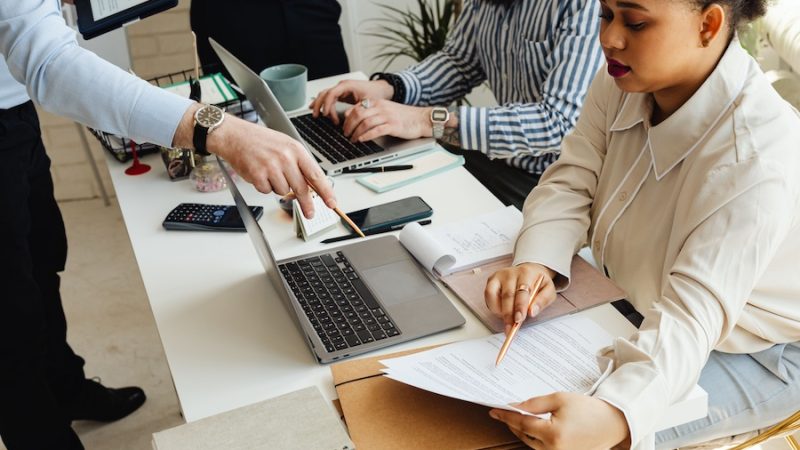 A busy office with two people working at laptops and a manager pointing to an important document.
